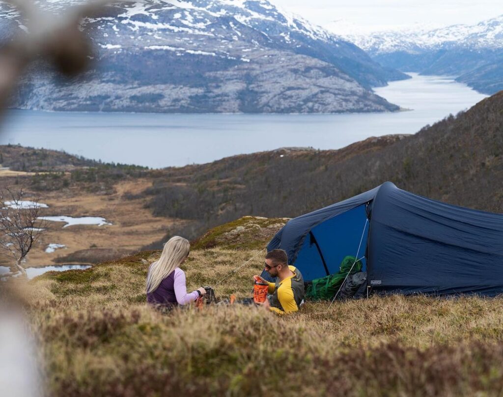 Une femme blonde et un homme assis sur l'herbe en face d'un lac et à côté d'une tente.