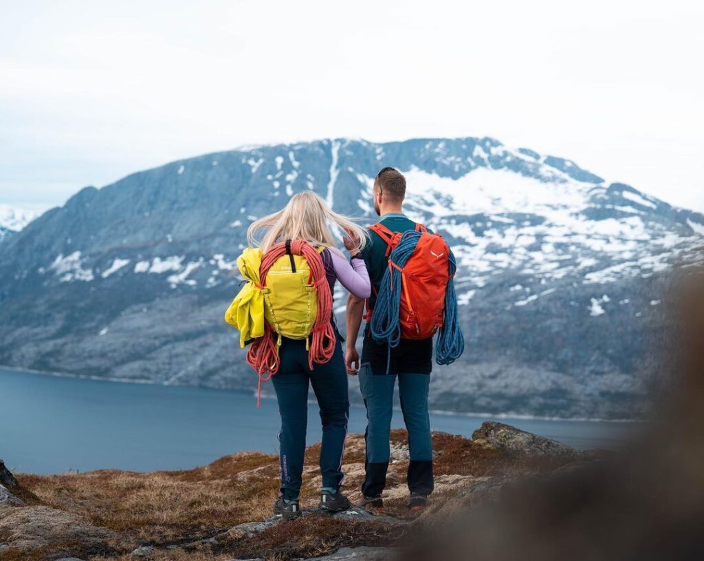 Un homme et une femme de dos devant une montagne et un lac, portant des cordes sur leurs sacs orange et jaune.